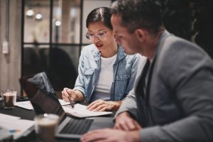 Two,Diverse,Businesspeople,Going,Over,Paperwork,Together,While,Working,At