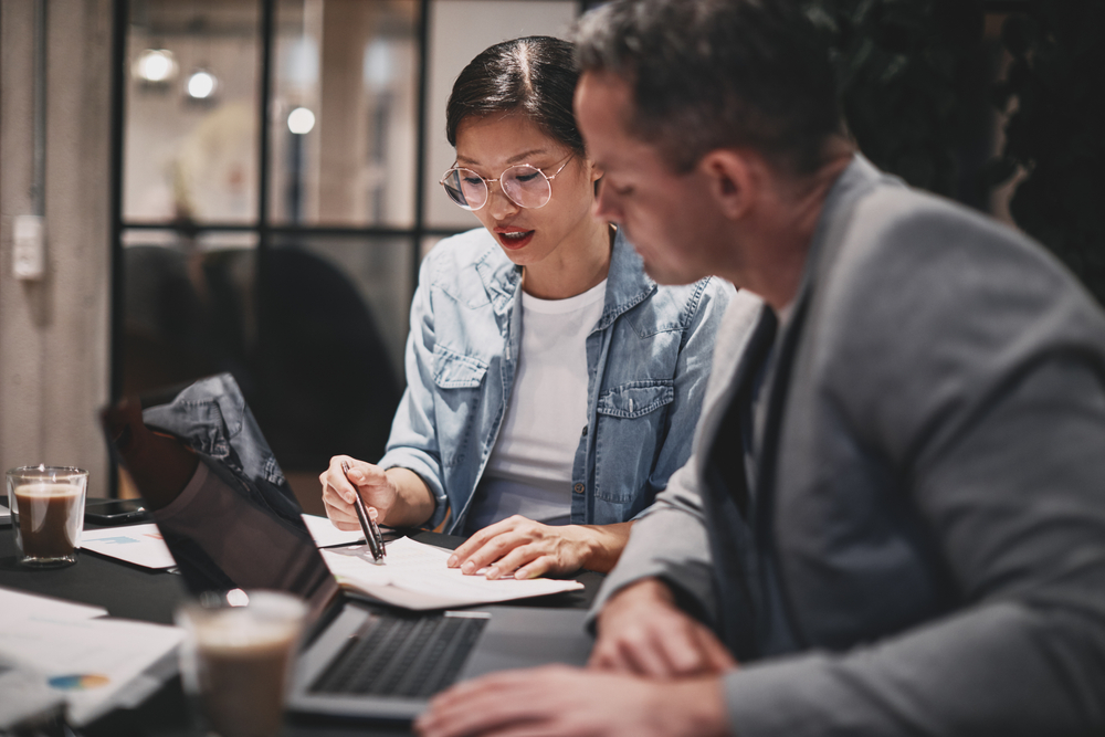 Two,Diverse,Businesspeople,Going,Over,Paperwork,Together,While,Working,At